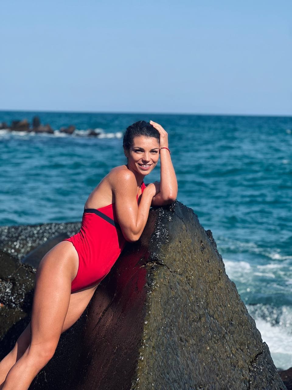 Полина в червен бански на скала край морето, усмихната